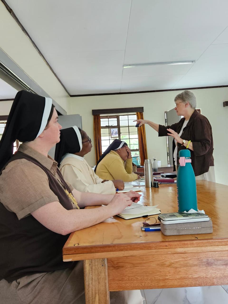 Three sisters sit at a table in a classroom listing to an older sister who stands before them.