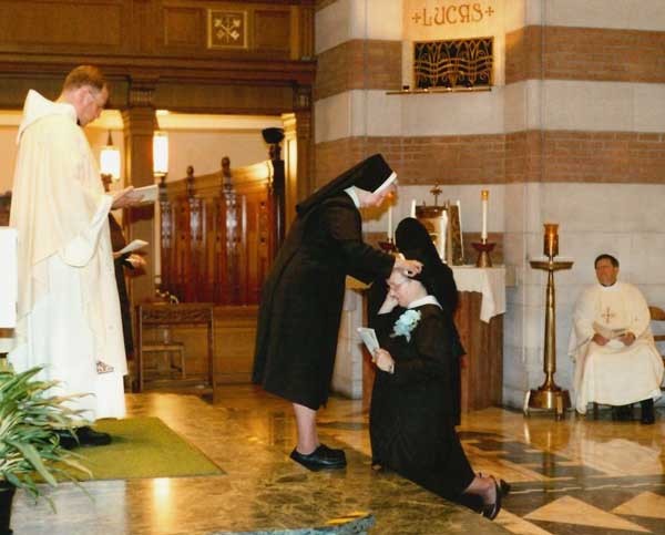 A sister is placing a viel on a kneeling sister's head near the alter in a chapel. A priest stands behind them on the alter and another priest is seated and watching from the ground floor.