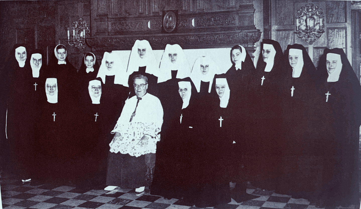 A group picture of novices, postulants, older sisters, and a priest, with some of them standing and sitting in front of a fireplace.