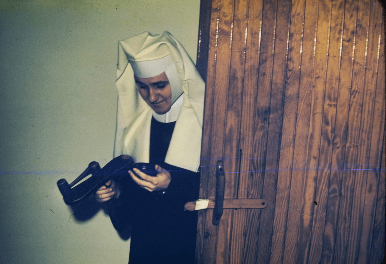 A novice standing in a doorway looking down at the clock in her one hand while a wooden clapper is in the other.