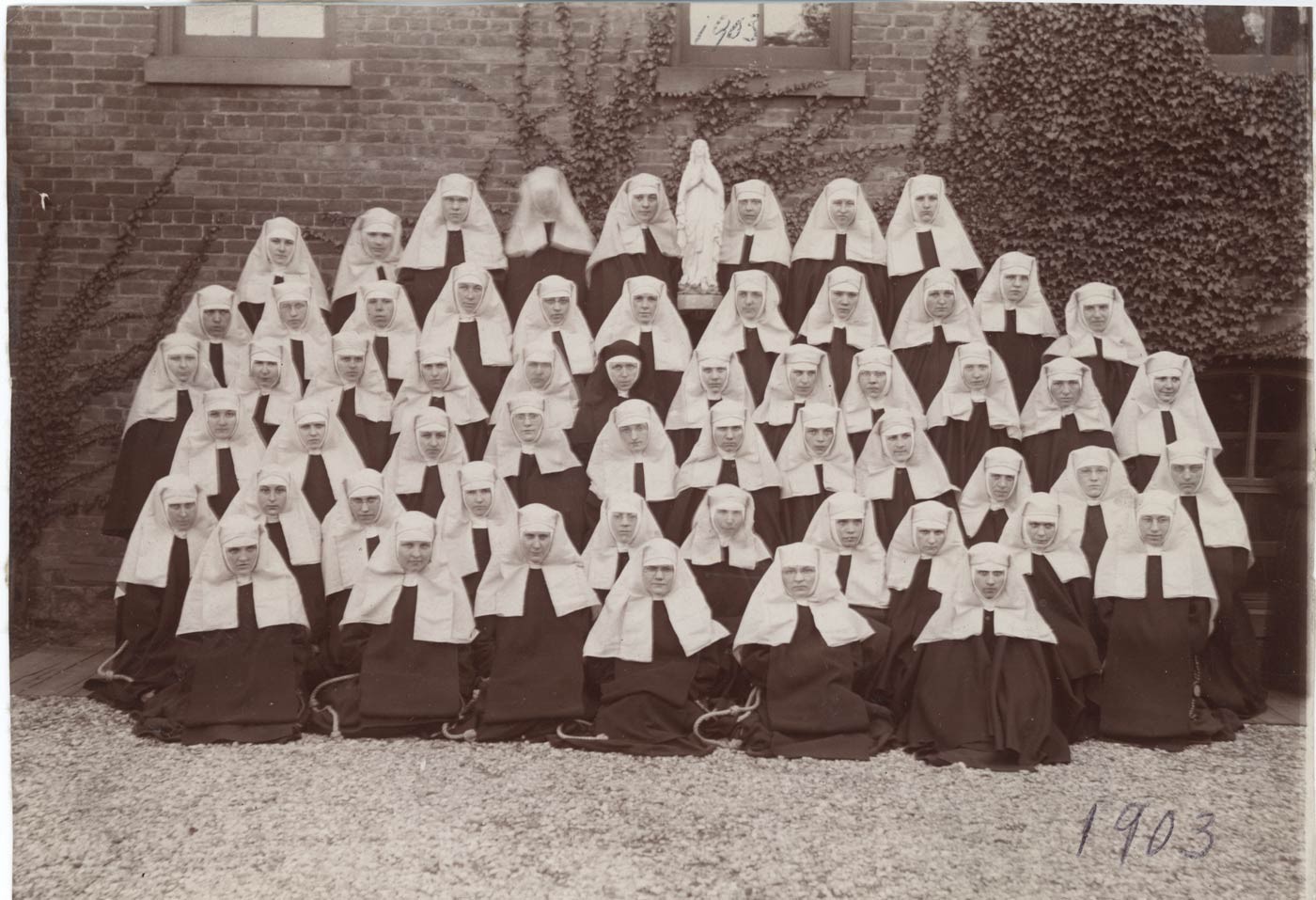 A large group of novices in several rows with one older sister situated in the center of them.