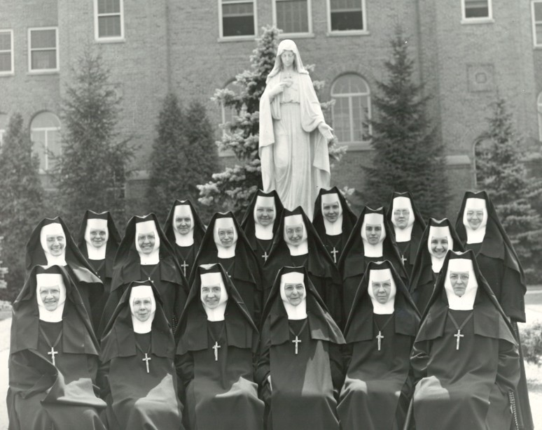 Group of sisters standing and sitting in front of a statue of the Blessed Mother.
